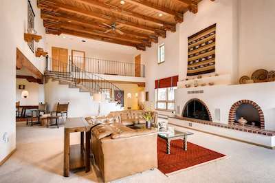The interior of a Santa Fe home with a family room featuring a fireplace, white walls, and a beamed ceiling. A staircase leads to a second floor interior balcony.
