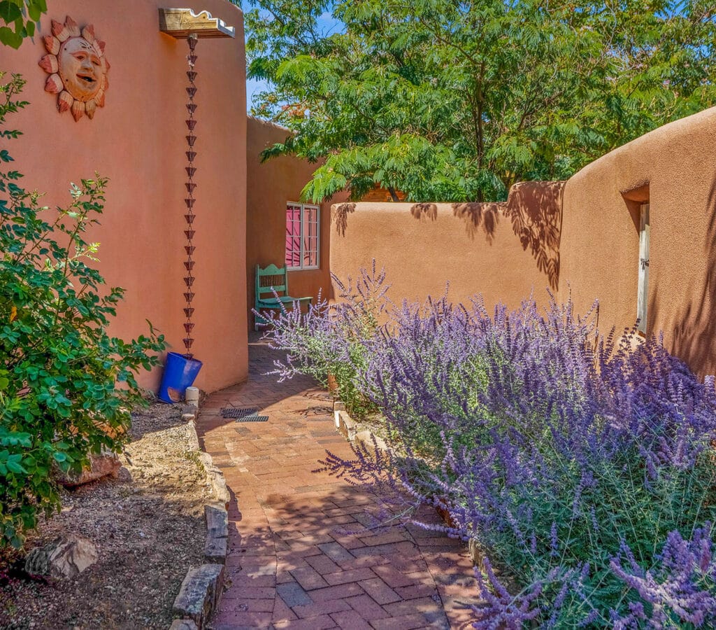 Walled courtyard with stucco walls. Brick pathway, canale with rain chain, colorful purple Russian sage plants and turquoise bench. Santa Fe, New Mexico.