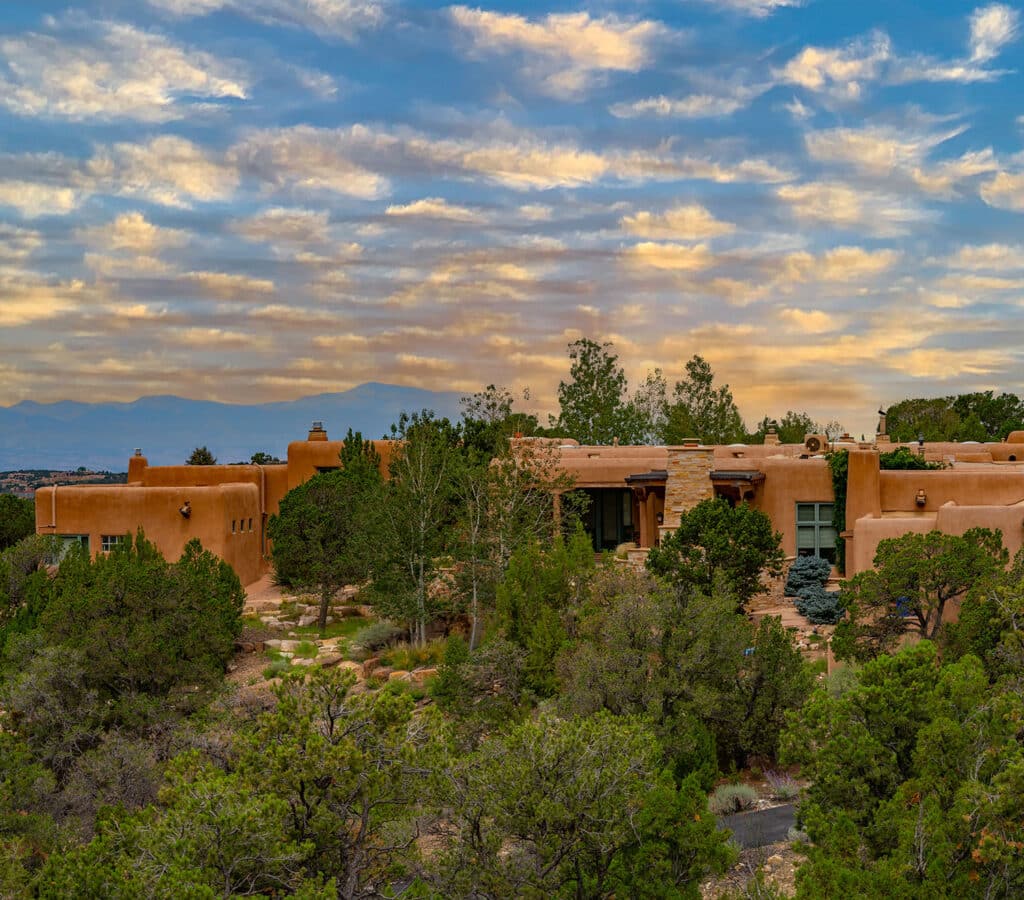 Single level Santa Fe style homes surrounded by trees with sunset clouds in the background.