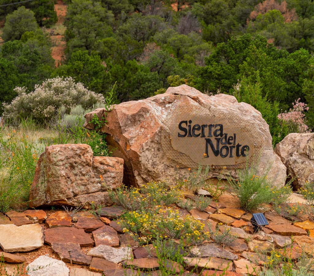 Stone boulders with community sign for Sierra del Norte in Santa Fe, NM. Green piñon trees in the background.