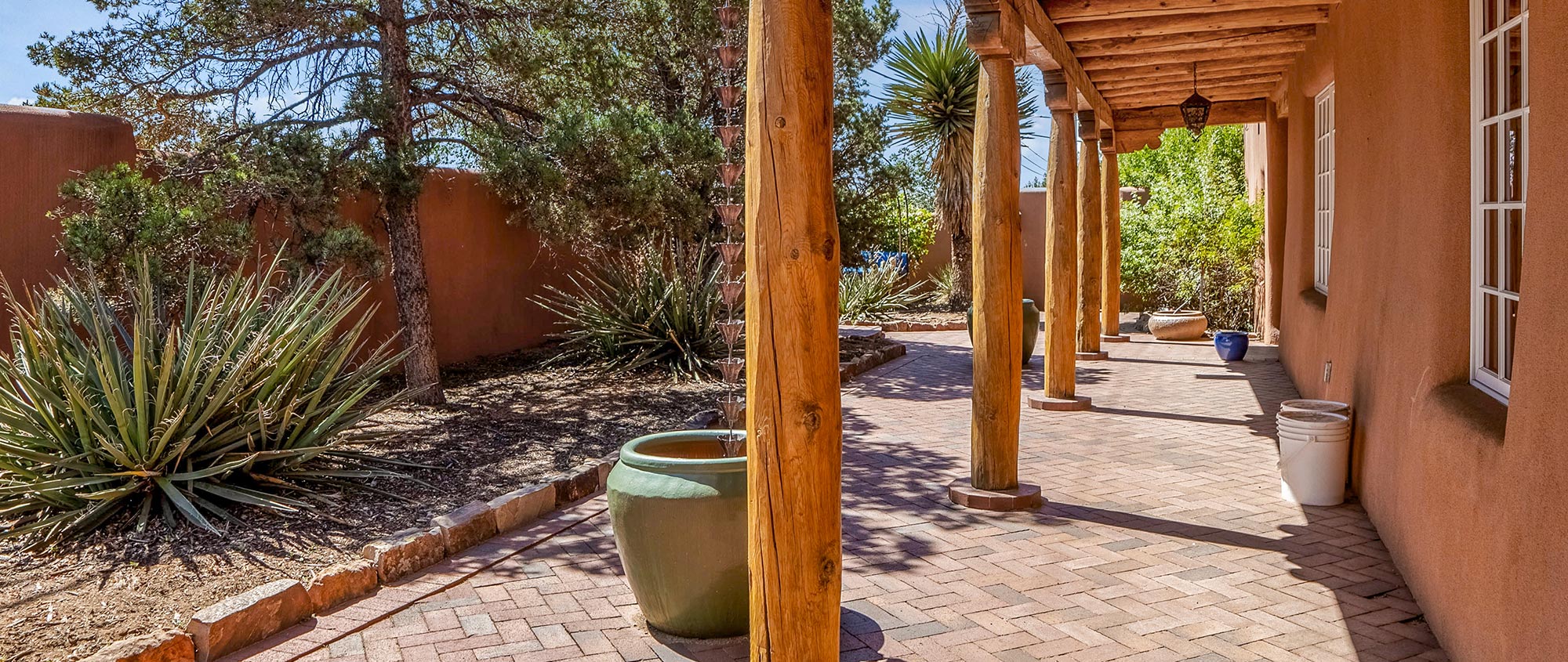 A covered portal with supporting posts, exterior bricks in walled courtyard with trees and yucca plants in Santa Fe, NM.